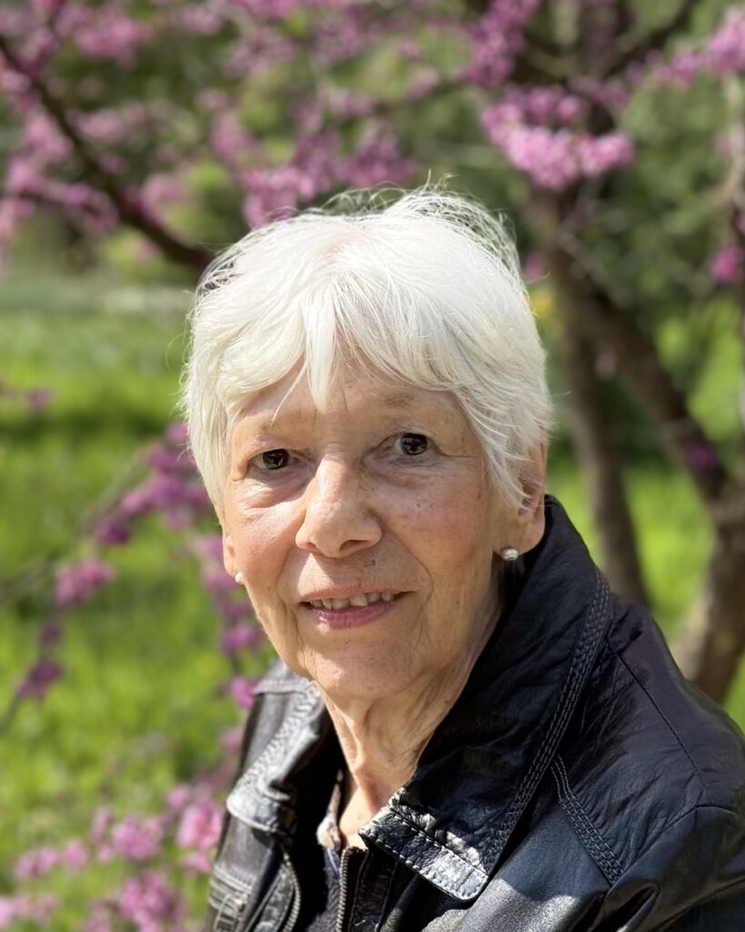 Porträt von Eva Boström mit weißem Haar und dunkler Lederjacke vor einem blühenden Judasbaum